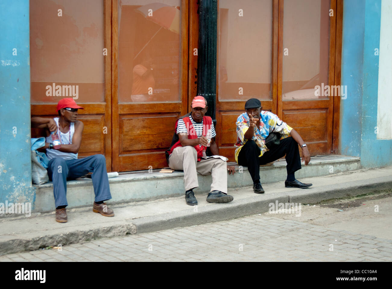 Three Cuban man are resting in a shade on the pavement, old Havana, Cuba one man is pointing at the camera Stock Photo