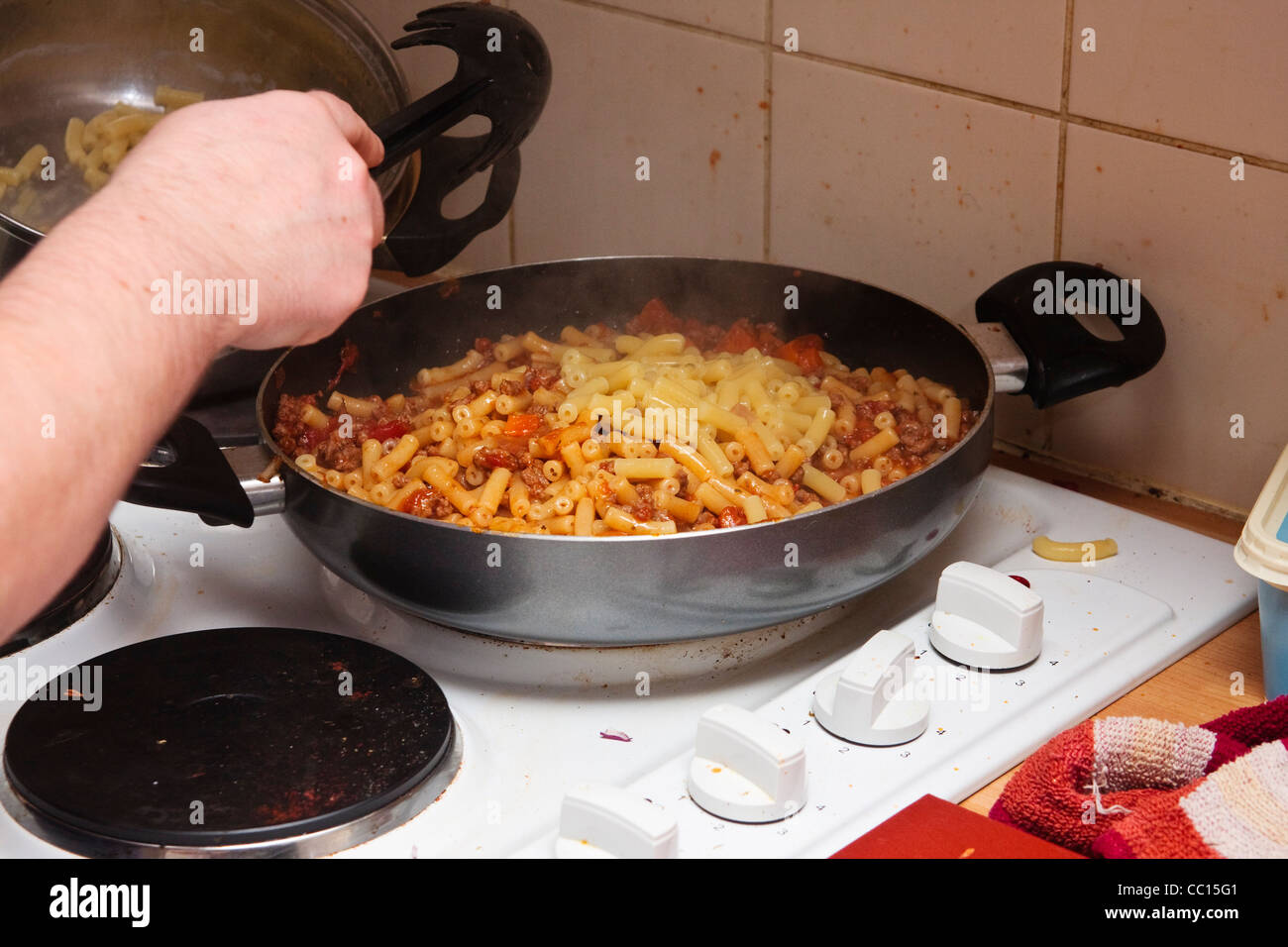 A female pours cooked macaroni pasta into a pan of bolognese Stock Photo