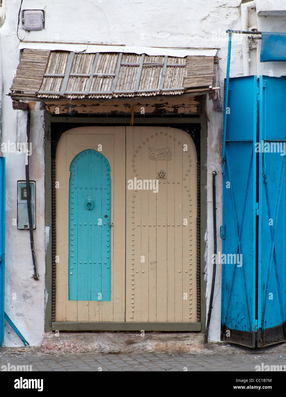A cream-painted wooden double door with an Arabian design and an inset smaller blue-painted door under a bamboo porch. Stock Photo