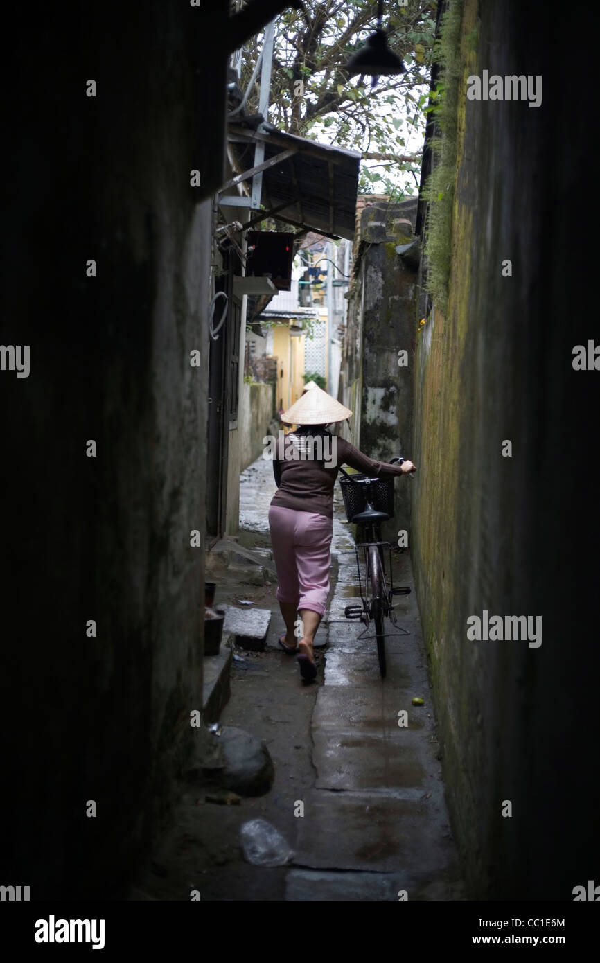 A woman in a conical hat walks down a narrow lane with her bicycle in Hoi An market, Vietnam Stock Photo