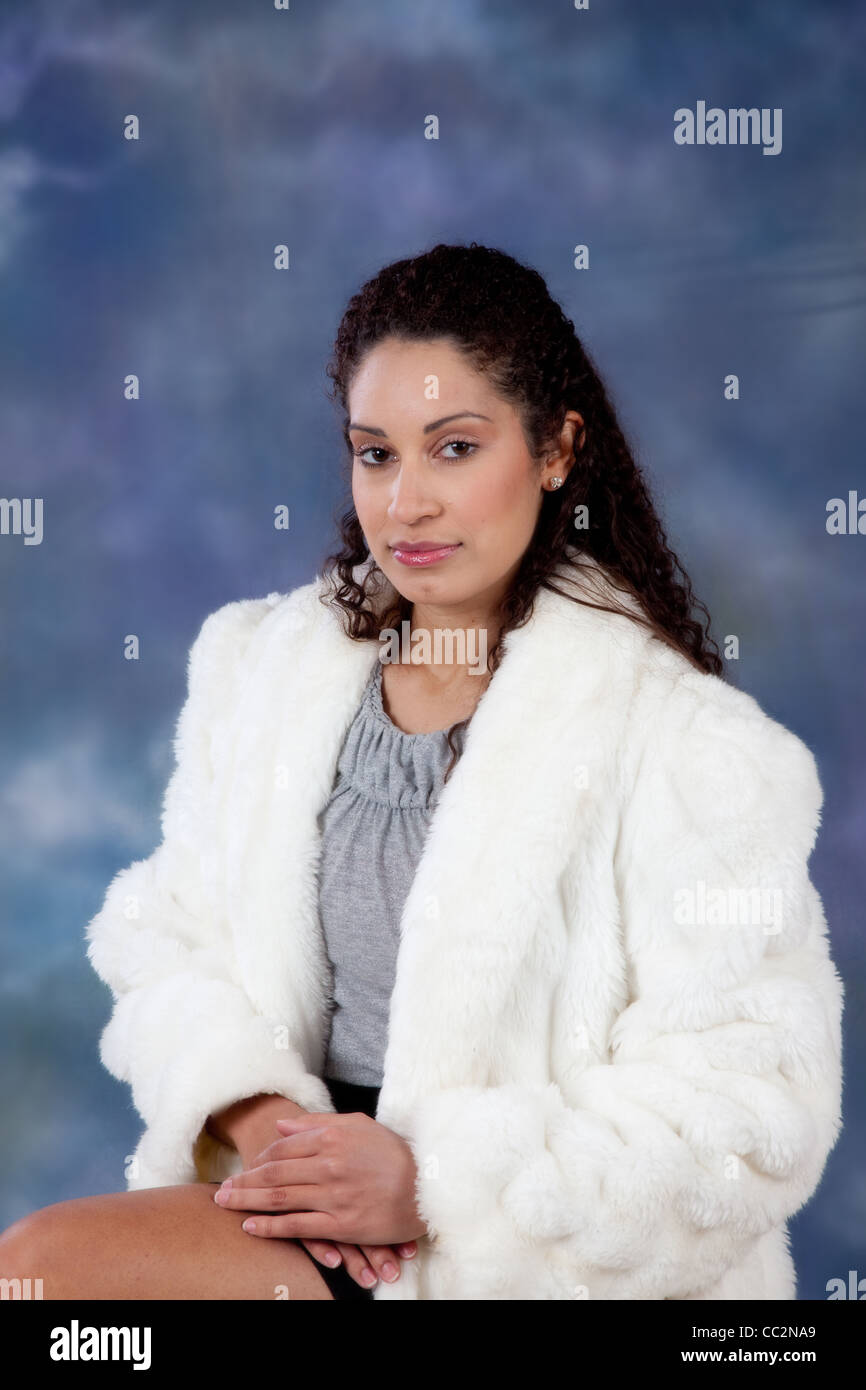 Happy and friendly woman in a white fur coat, sitting with eye contact and a slight smile for the camera Stock Photo