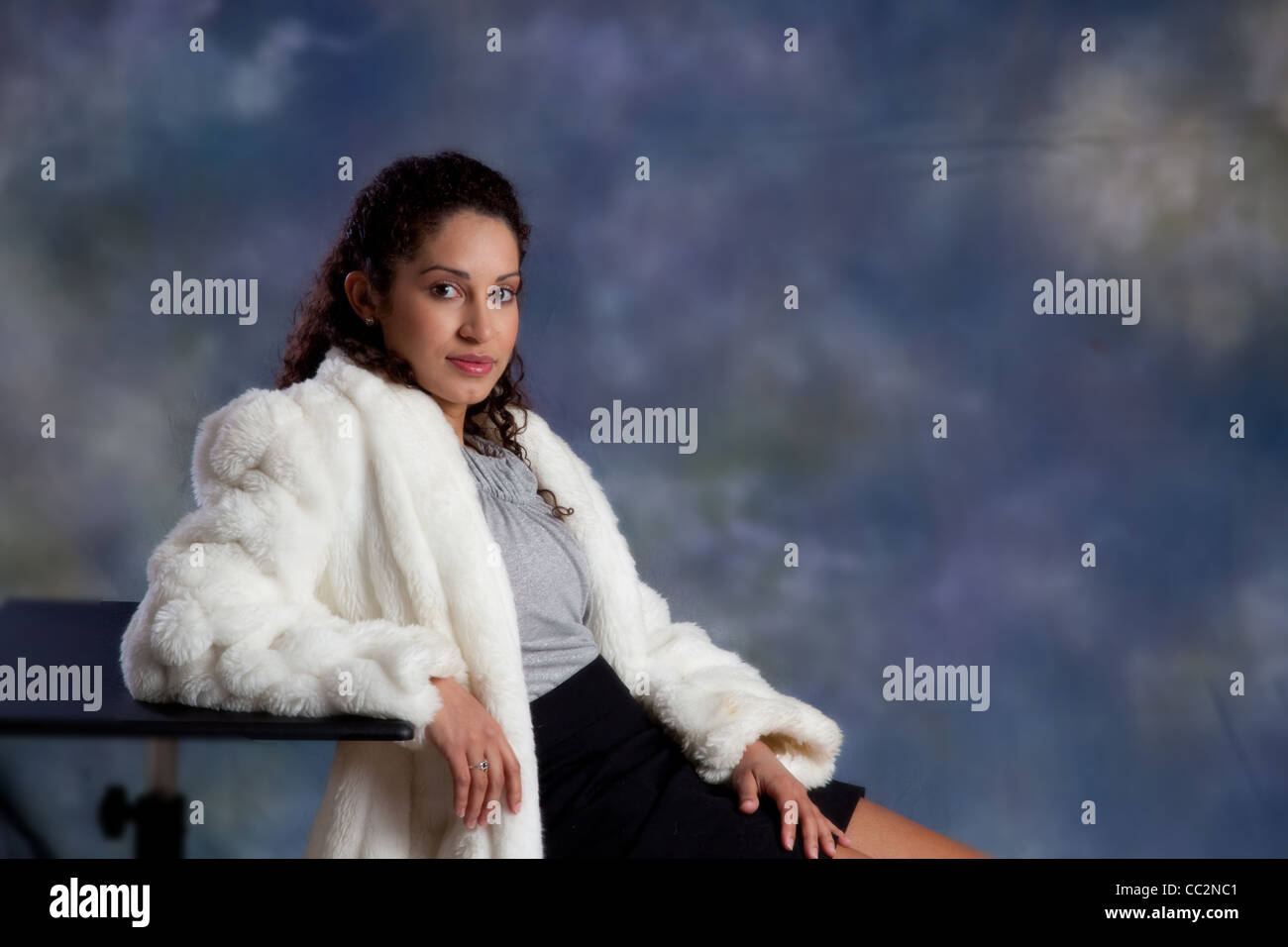 Happy and friendly woman in a white fur coat, sitting with eye contact and a slight smile for the camera Stock Photo