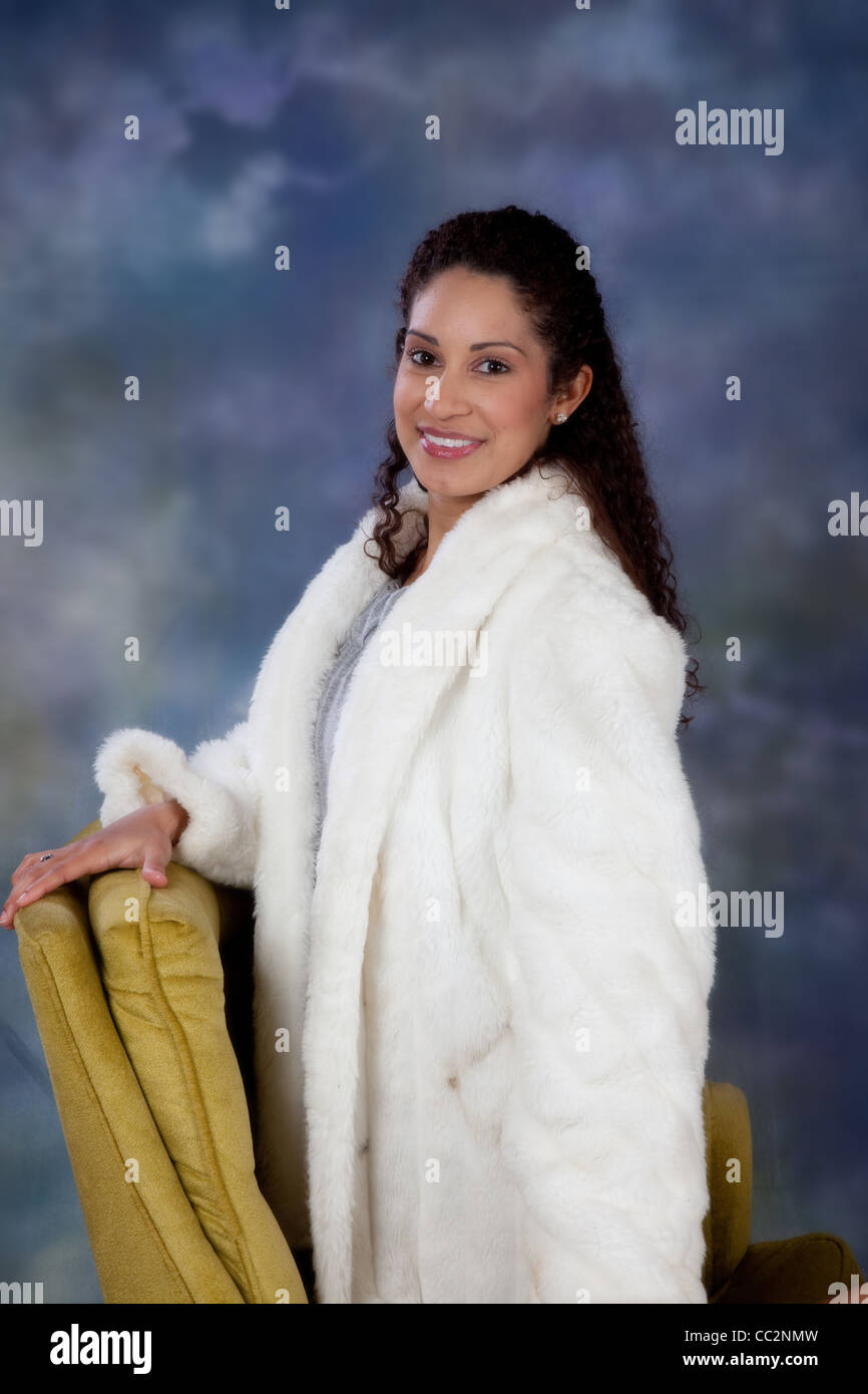 Happy and friendly woman in a white fur coat, kneeling in a chair and looking at the camera with a pleased smile Stock Photo