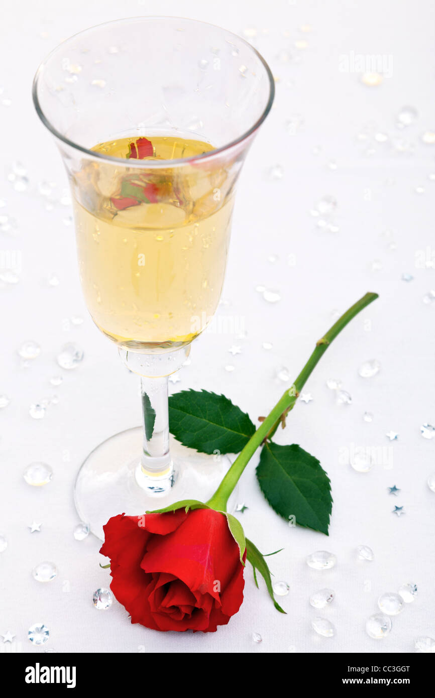 Photo of a single red rose and a glass of champagne with fake diamond table confetti. Focus is on the rose. Stock Photo