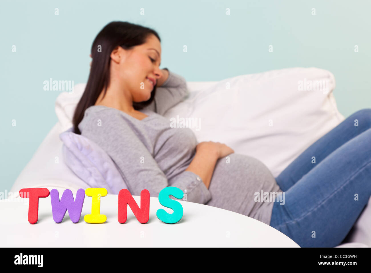Photo of a pregnant woman at home sitting in an armchair, focus on the word TWINS on the table in front. Stock Photo