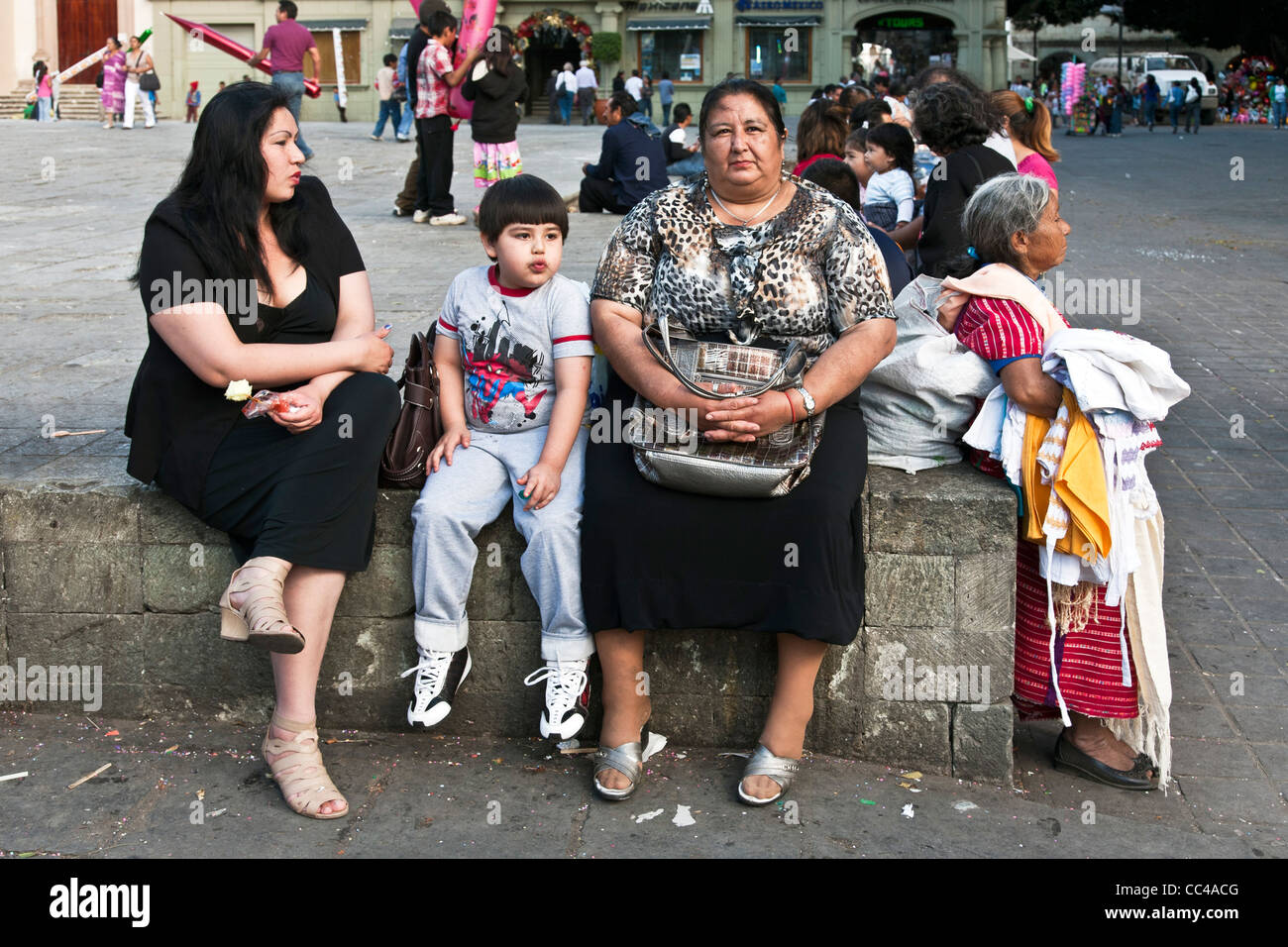 buxom Mexican grandmother with plump daughter & chubby young grandson sitting on a wall at edge of Zocalo next to Triqui vendor Stock Photo