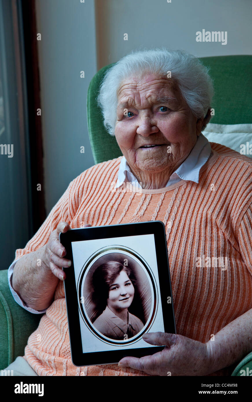 98 years old elderly lady holding an iPad tablet computer, displaying a sepia portrait of herself taken 80 years ago, at age 18 Stock Photo