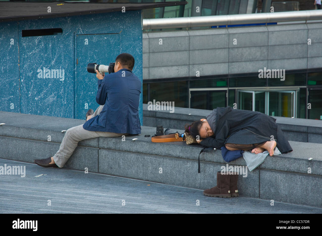 Asian man with long zoom lens taking photographs while partner is taking a nap London England UK Europe Stock Photo