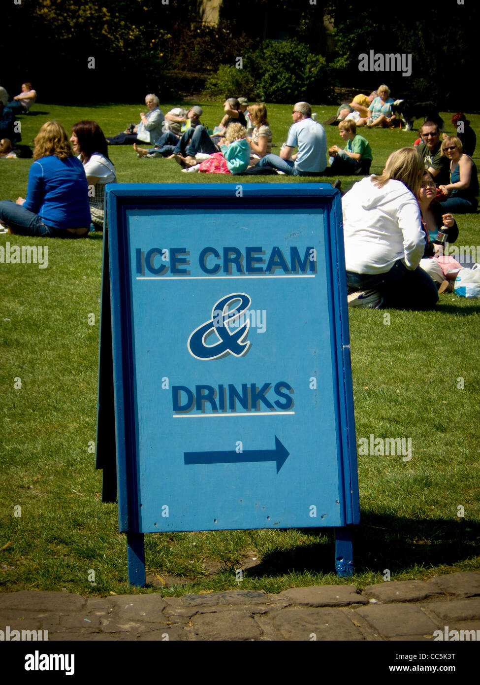 People sitting on the grass in a public green space, in summer, with an A-board in the foreground with directions for ice cream and drinks. Stock Photo