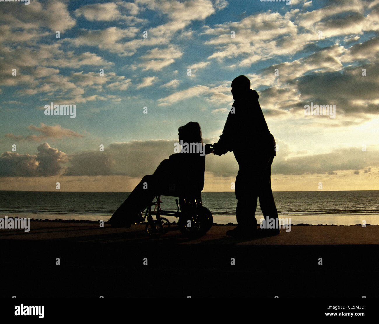 Silhouetted male pushing an unidentifiable person in a wheelchair along seafront as the sun sets. UK Stock Photo
