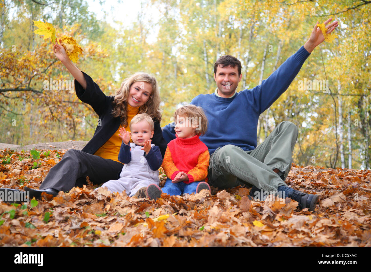 family of four sits in autumn park. parent hands up. Stock Photo