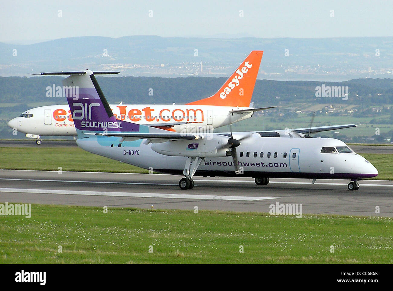 Two aircraft at Bristol International Airport, Bristol, England. The orange easyJet queueing for take off is Boeing 737-700 G-EZJW. The propeller aircraft starting take off is Air Southwest de Havilland Dash 8, G-WOWC. Stock Photo