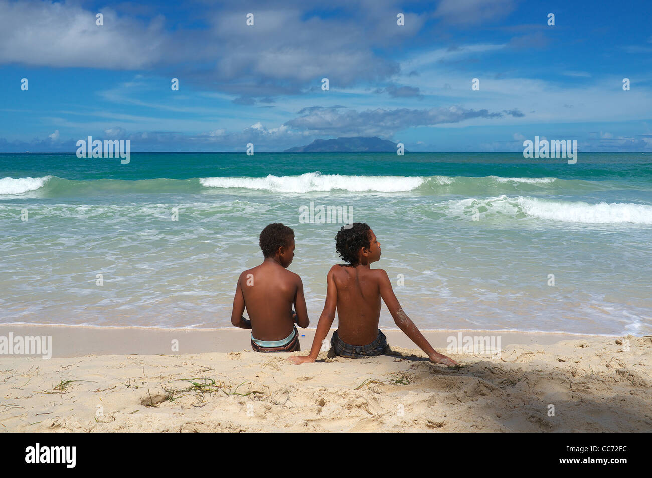 Two young boys sitting on the sand at a beach in Mahe, Seychelles, at the Indian Ocean with Silhouette Island on the Horizon Stock Photo