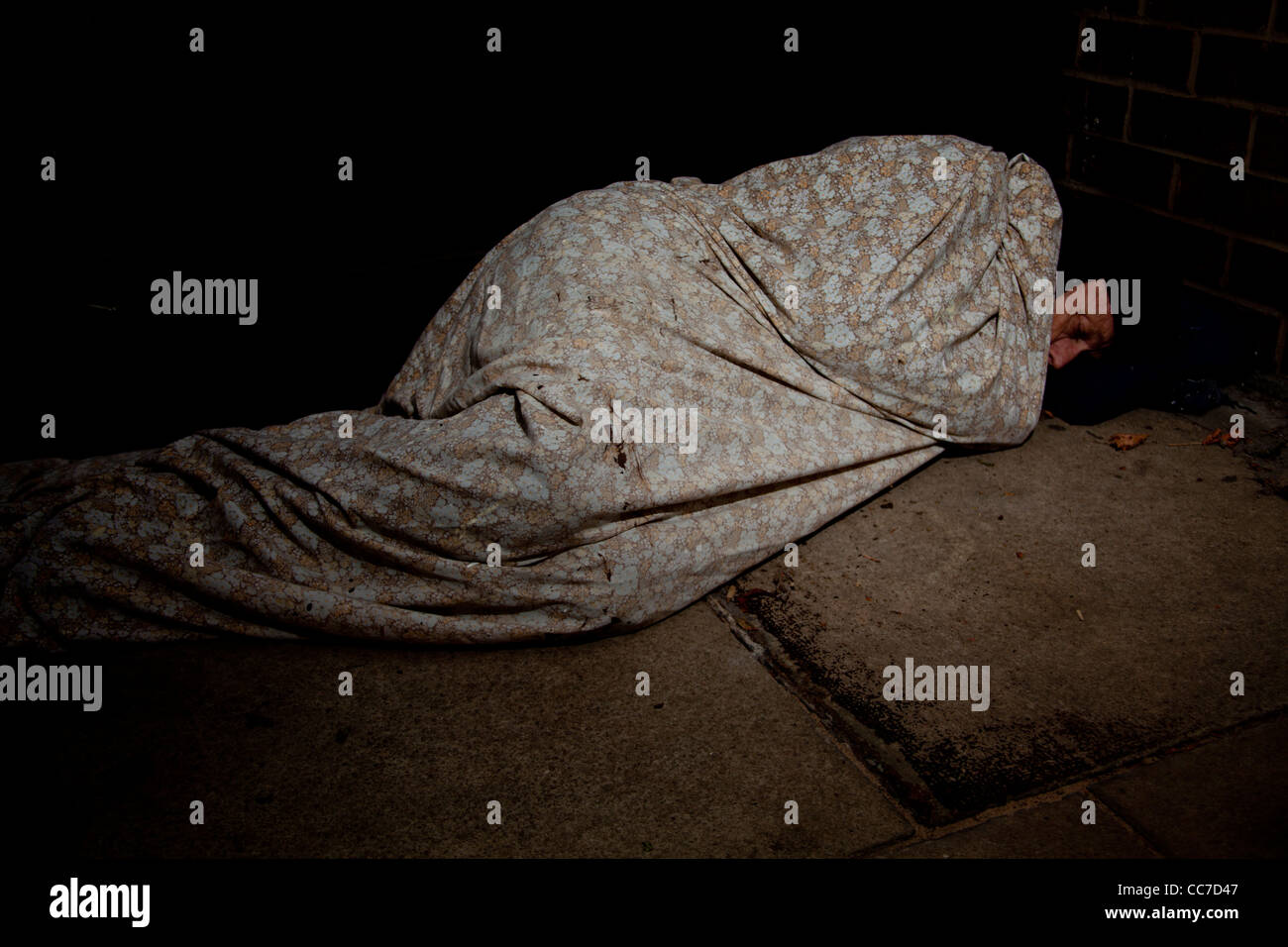 Image depicting a homeless man sleeping covered in a blanket on the cold stone pavement. Stock Photo