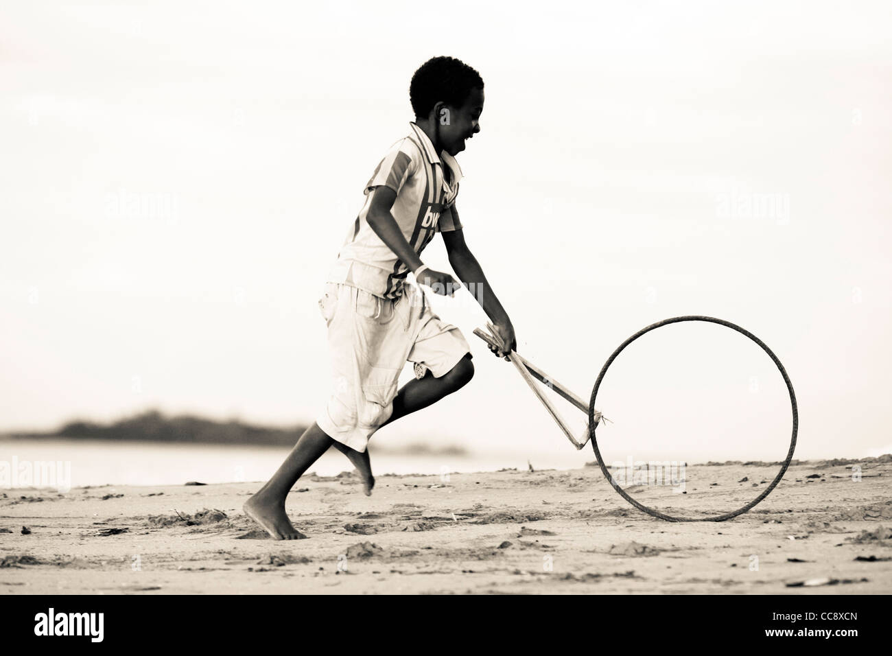 A young malagasy boy 6-10 years running with a stick and a bicycle rim, a traditional toy, at the beach. Nosy Komba, Madagascar Stock Photo