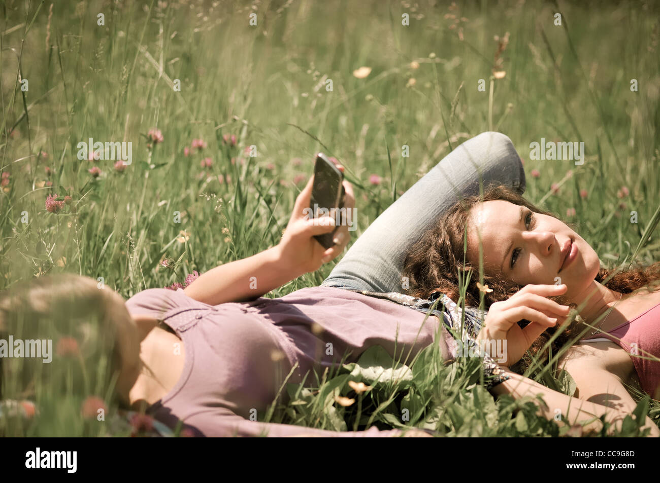 Two friends (teenage girls) relax lying on grass outside in nature - one girl is using cellphone Stock Photo