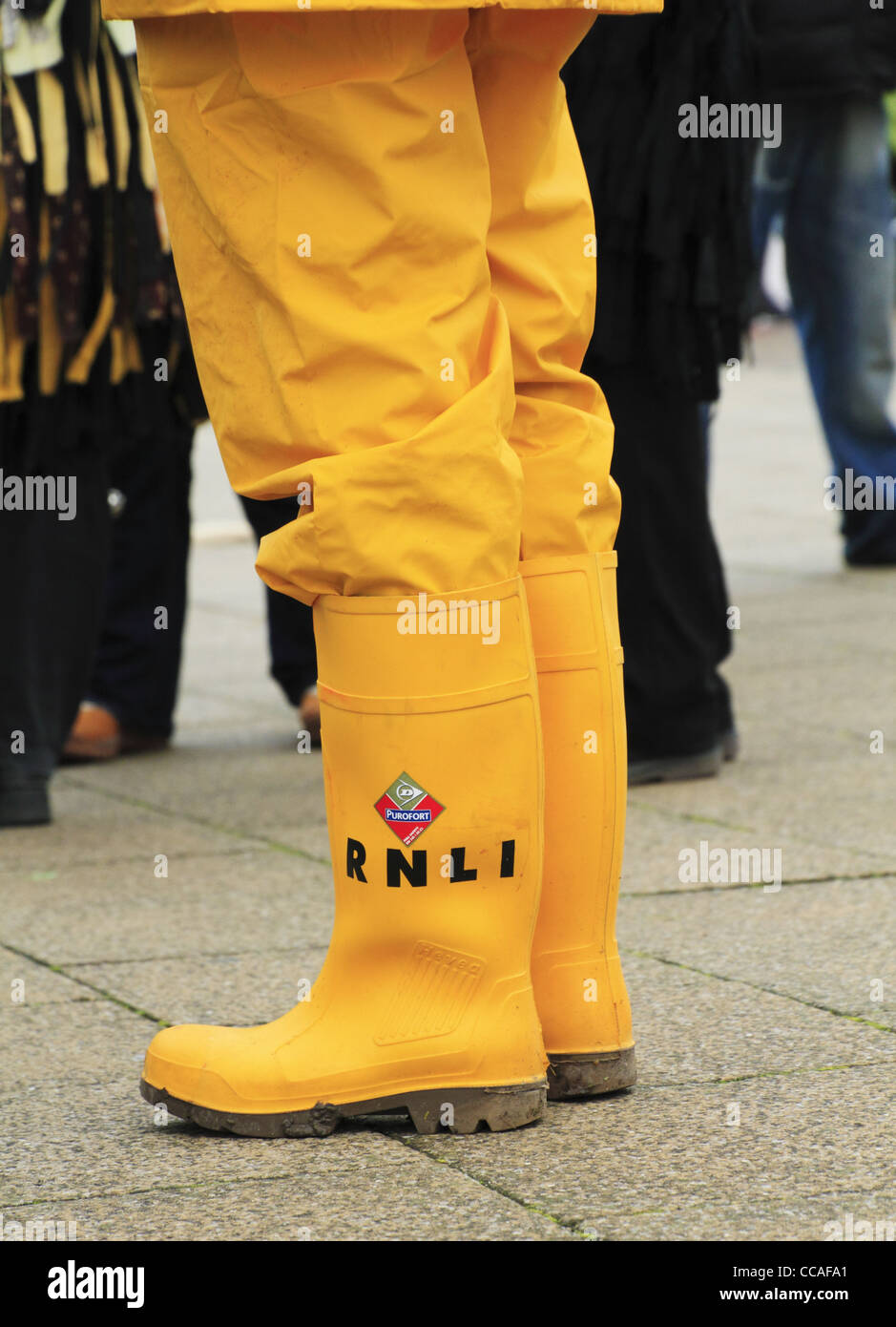 An RNLI Lifeboats volunteer in bright yellow sou'wester uniform wet weather gear wellies wellington boots Eastbourne Seafront. Stock Photo