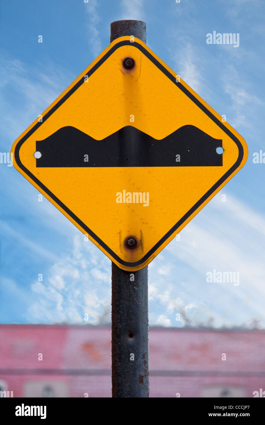 A bumpy road ahead sign on a rusty pole against a blue sky with wight clouds and a red building Stock Photo