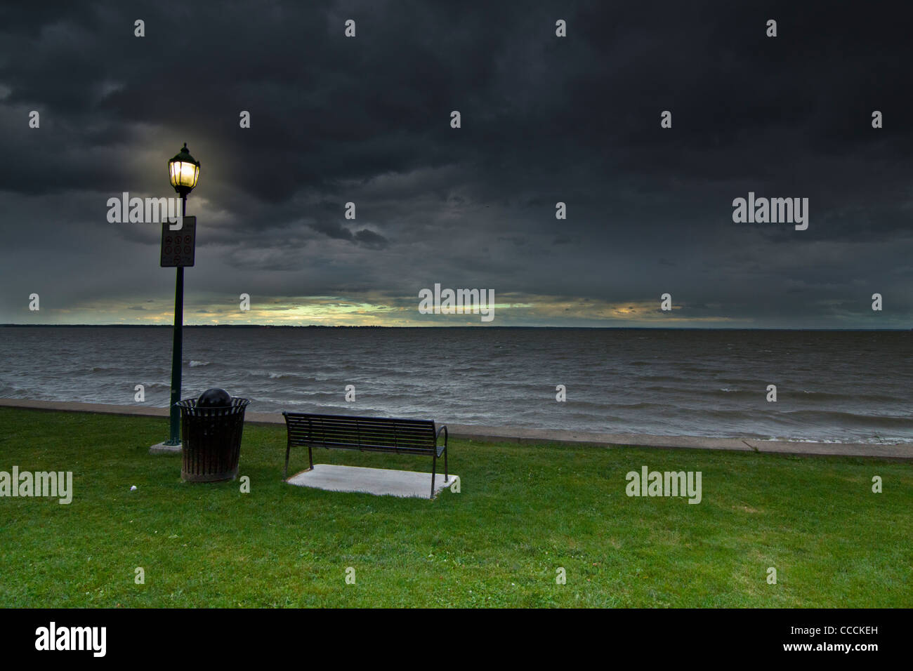 A public lighting lantern pole near a metal bench and a bin, on green grass, near the river side under a heavy stormy sky Stock Photo