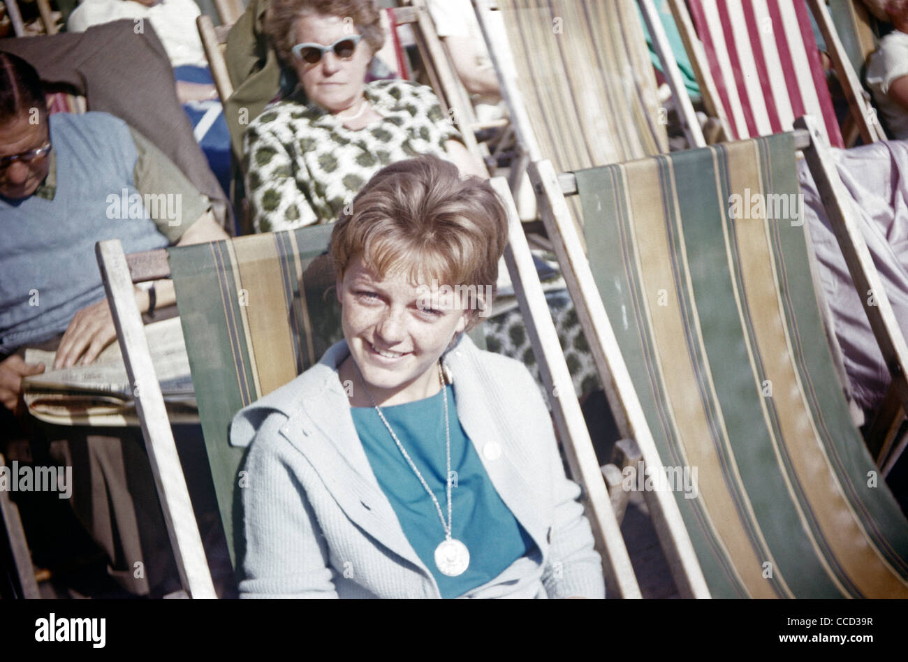 A teenage girl smiles in summer sunshine while sitting on a seafront deckchair in the early nineteen sixties. Stock Photo