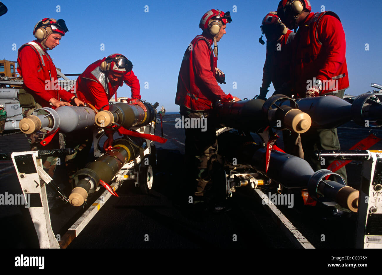 Red shirted ordnance men prepare to fit smart bombs and missiles to an F/A-18 fighter jet on deck of USS Harry S Truman. Stock Photo