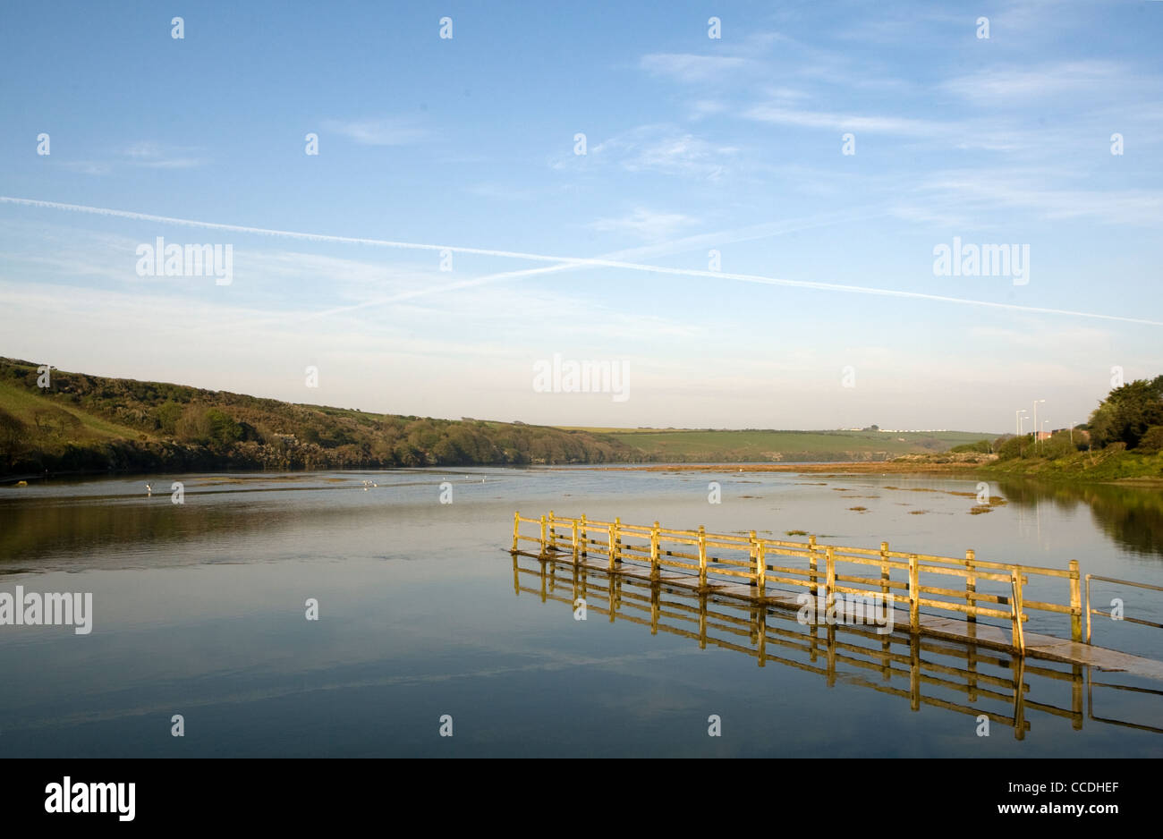Vapour trails cross over a walkway which is partially submerged by the incoming tide on the River Gannell at Newquay, Cornwall. Stock Photo