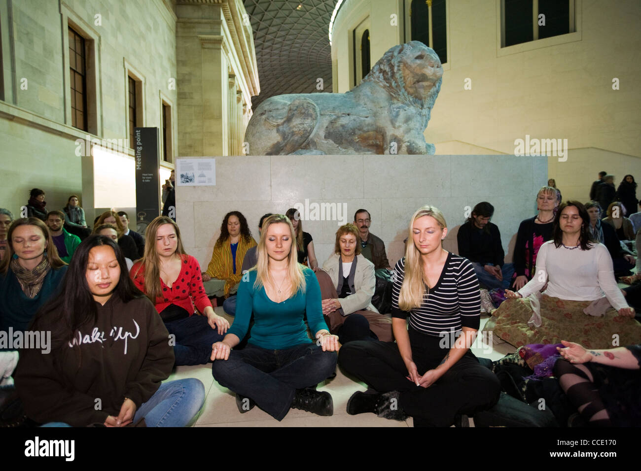 People meditate as part of a meditation flash mob in the Great Court of the British Museum beneath a stone lion statue Stock Photo