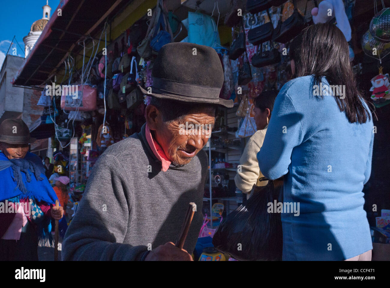 An elderly Ecuadorian man with a very wrinkled face wearing a fedora hat and holding a walking stick in marketplace. Stock Photo