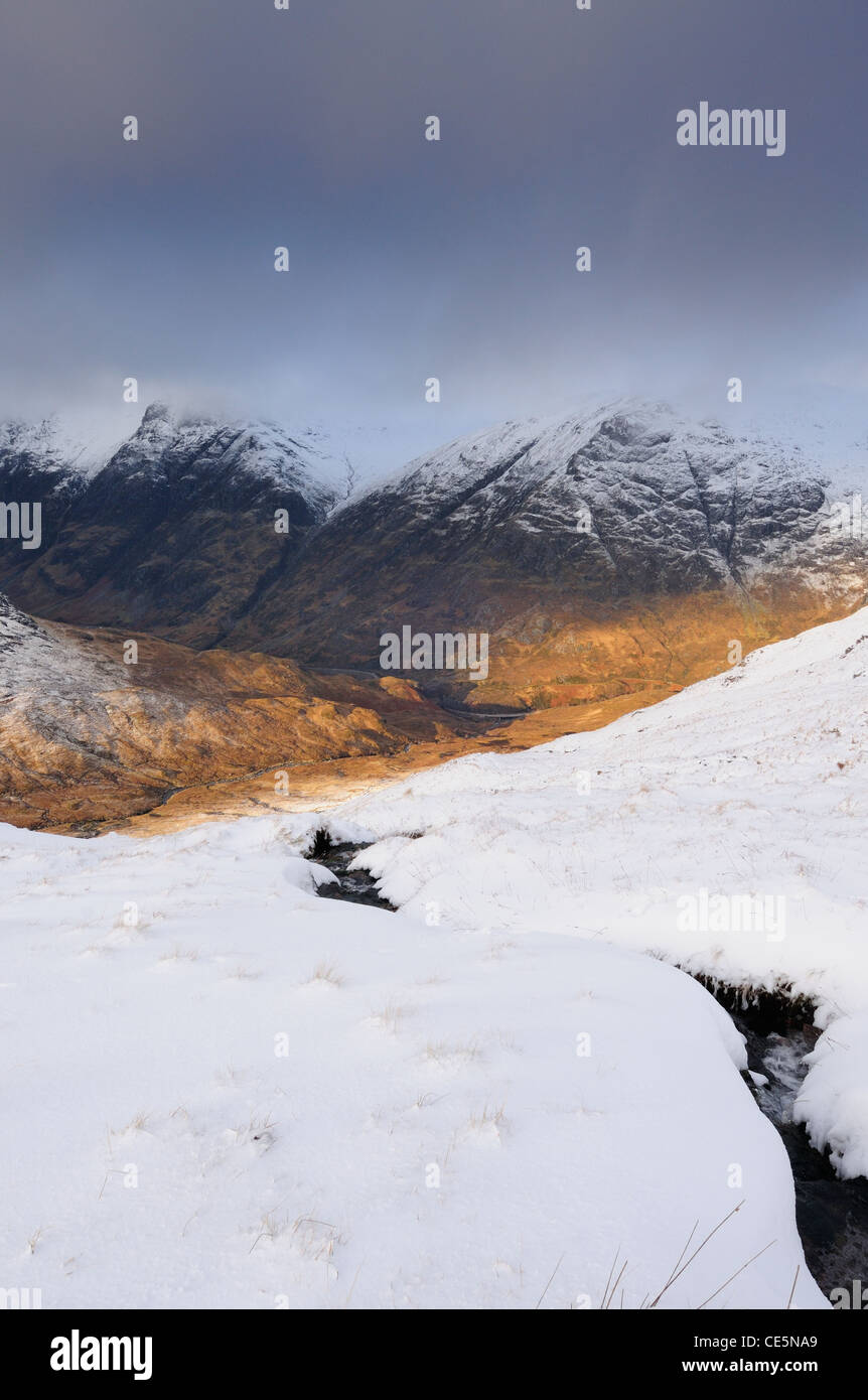 Stream through the snow on Buachaille Etive Beag, with sunlight on the Pass of Glencoe and Lairig Eilde Stock Photo