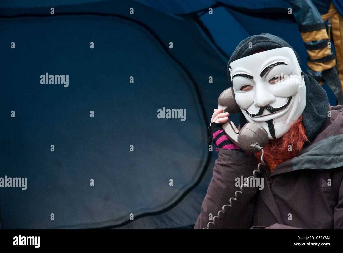 A masked protester wearing Guy Fawkes mask, camped at St Pauls Occupy London site on an old telephone, tent in background. Stock Photo