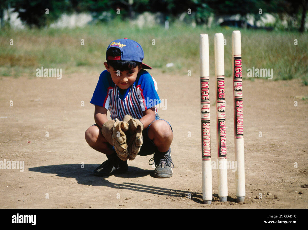 Indian child boy playing cricket doing wicket keeping sitting behind stumps ; India ; Asia ; MR#158 Stock Photo