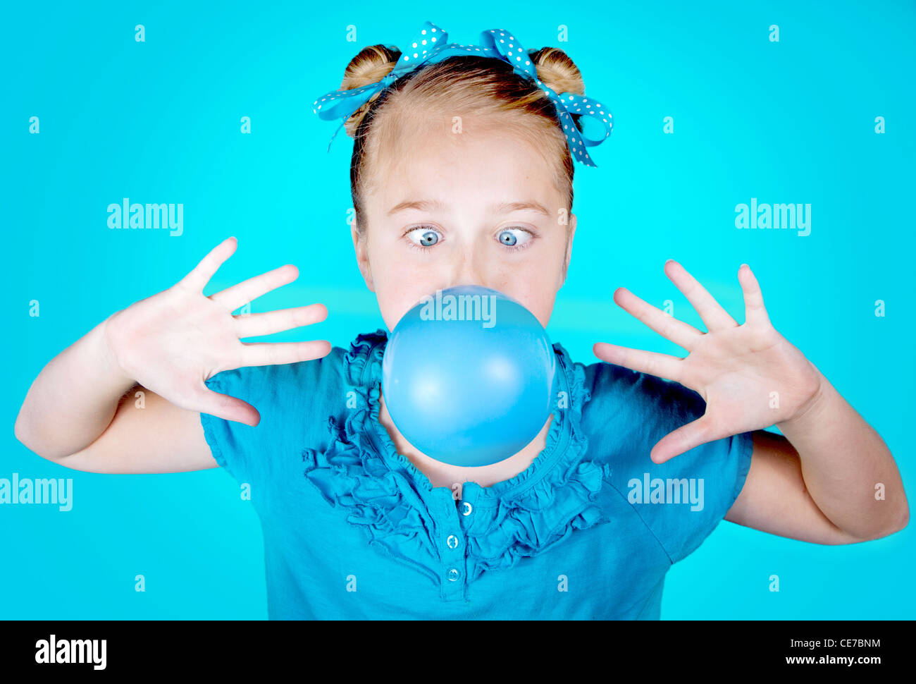 a cross eyed girl wearing blue with blue eyes and a blue bow, blowing a blue bubble. On a blue background Stock Photo
