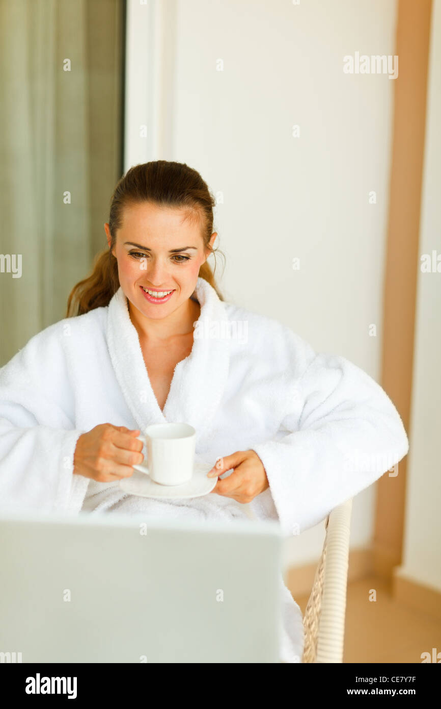 Happy woman in bathrobe sitting on terrace having coffee and exploring something in notebook Stock Photo