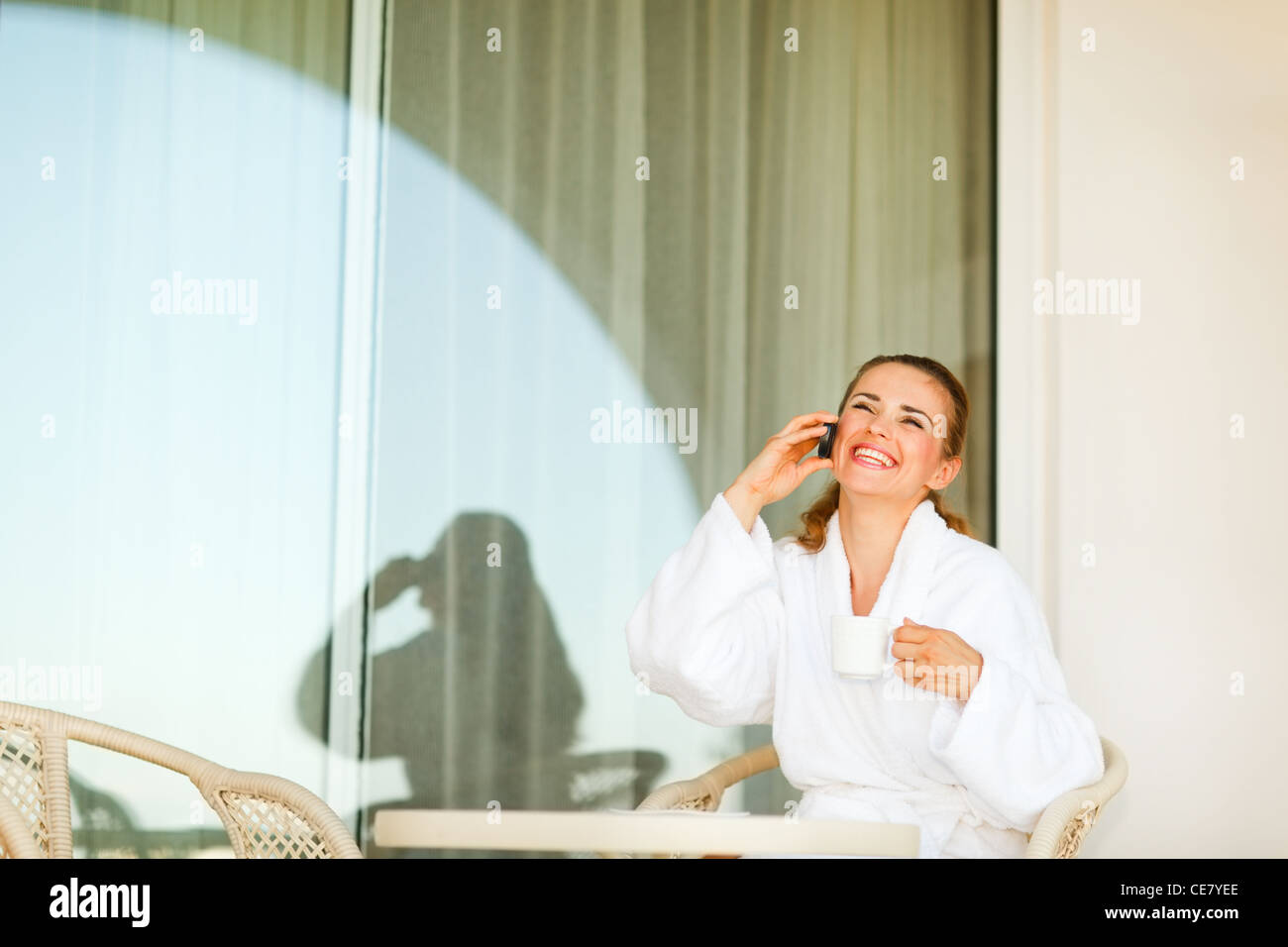 Laughing young woman in bathrobe with cup of coffee speaking mobile phone on terrace Stock Photo