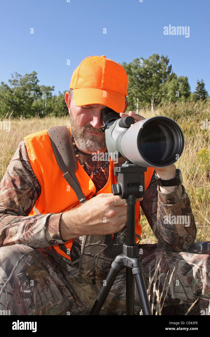 orange-clad hunter sitting and looking through spotting scope Stock Photo