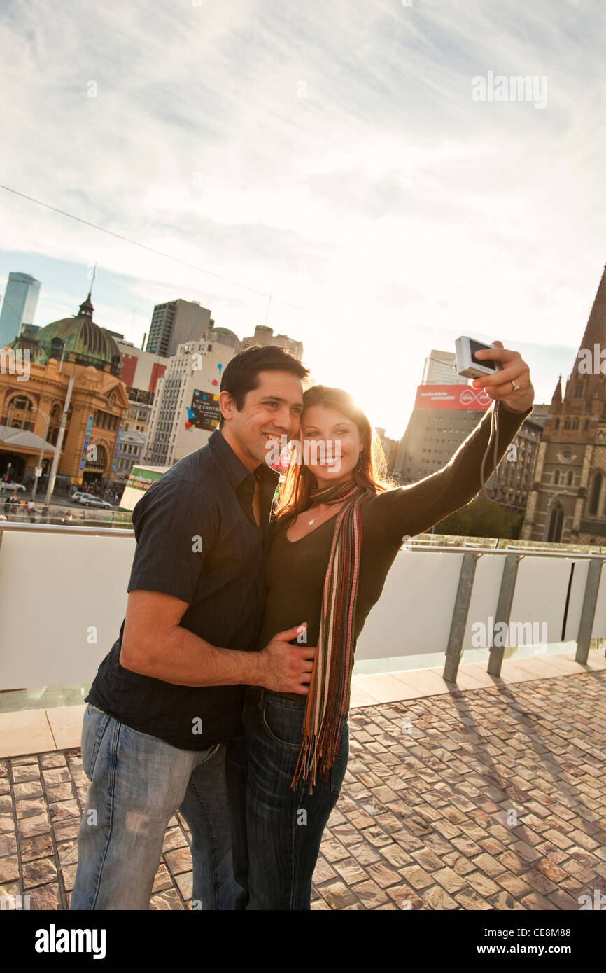 Couple taking a self-portrait with city skyline in background at sunset. Federation Square, Melbourne, Victoria, Australia Stock Photo