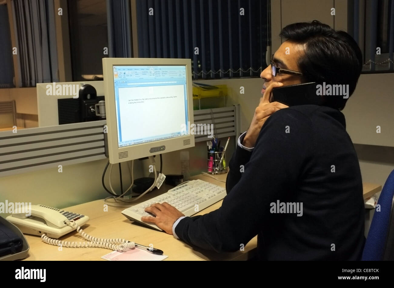 An office worker takes a call on his mobile phone, whilst working at computer Stock Photo
