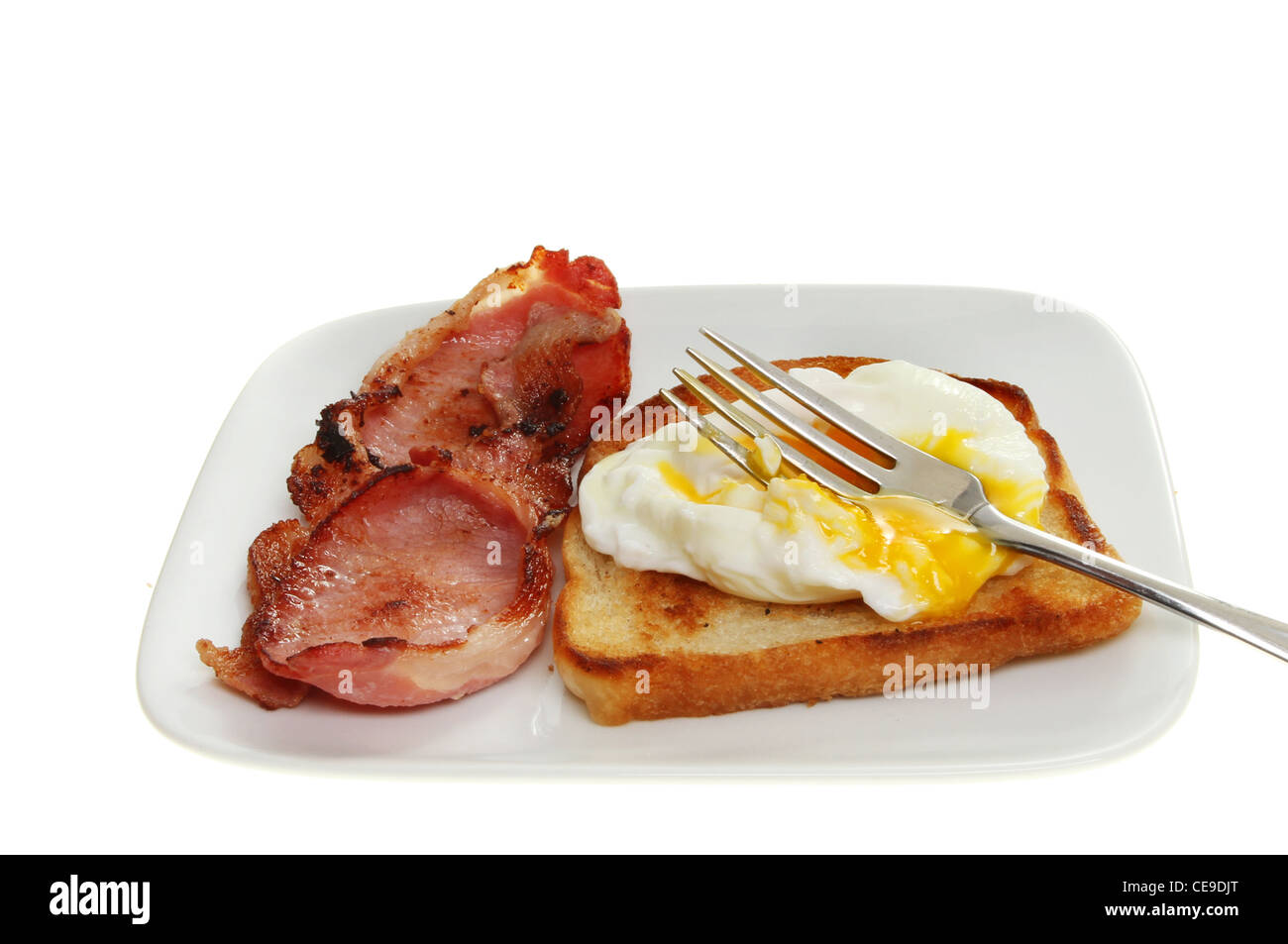 Poached egg with a fork resting in runny yolk with fried bread and bacon on a plate isolated against white Stock Photo