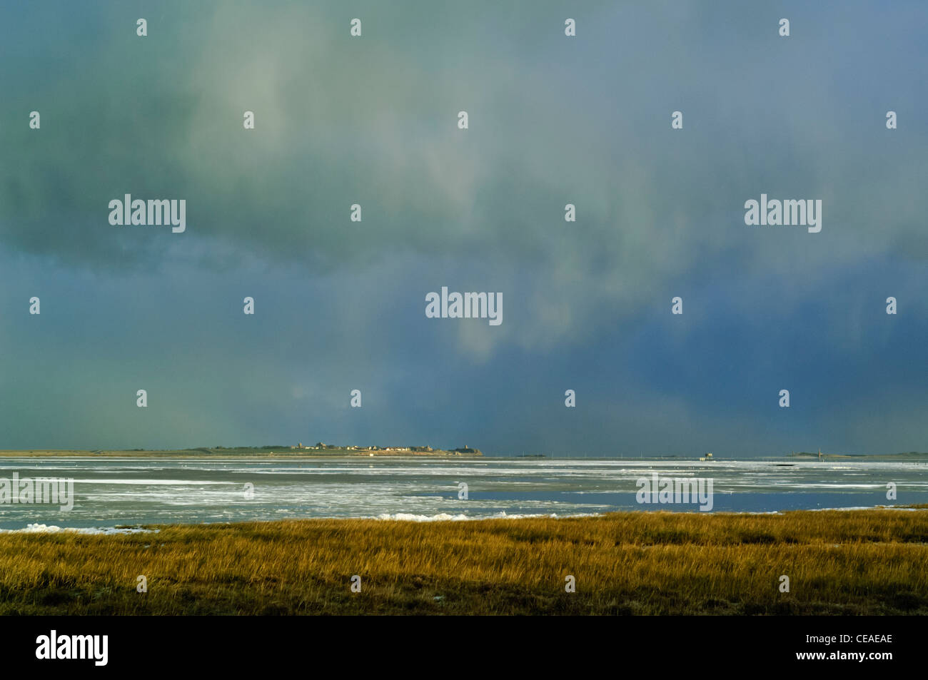 Lindisfarne Island (Holy Island) and village, beneath stormy sky and grey clouds, across icy water and frozen tidal mudflats Stock Photo