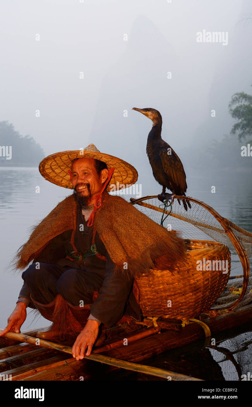 Smiling cormorant fisherman with bird on a net on the Li river at dawn with Karst mountain peaks Xingpingzhen Yangshuo, Guilin, Guangxi China Stock Photo