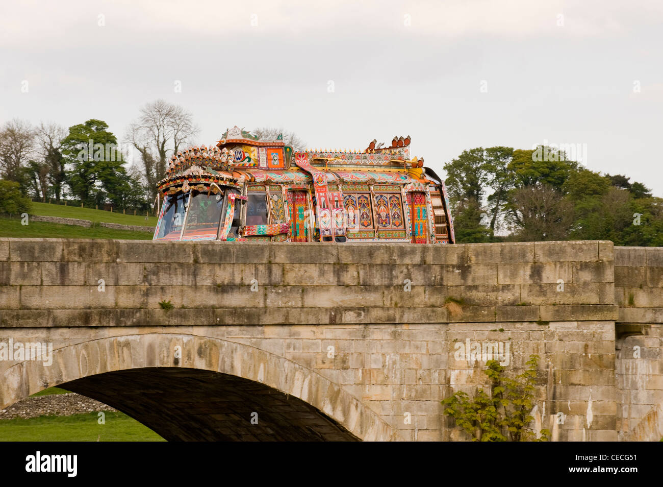 Colourful decorated Indian style bus driving over old bridge in Yorkshire Dales (on location, filming Sharwood's ad campaign) - Burnsall, England, UK. Stock Photo