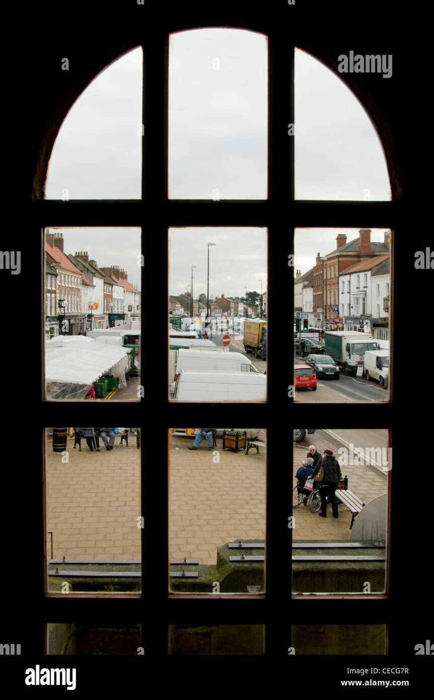 View through window of busy bustling town (Northallerton High Street) on market day (stalls, people shopping, road) - North Yorkshire, England, UK. Stock Photo