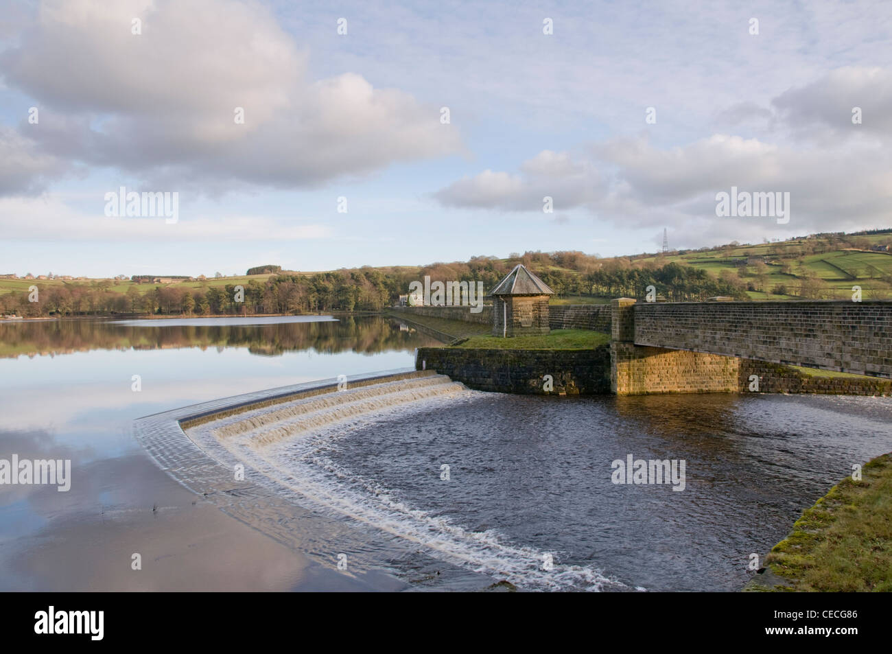 Water flowing over spillway from calm scenic tree-lined lake on sunny evening - Swinsty Reservoir, Washburn Valley, North Yorkshire, England, UK. Stock Photo