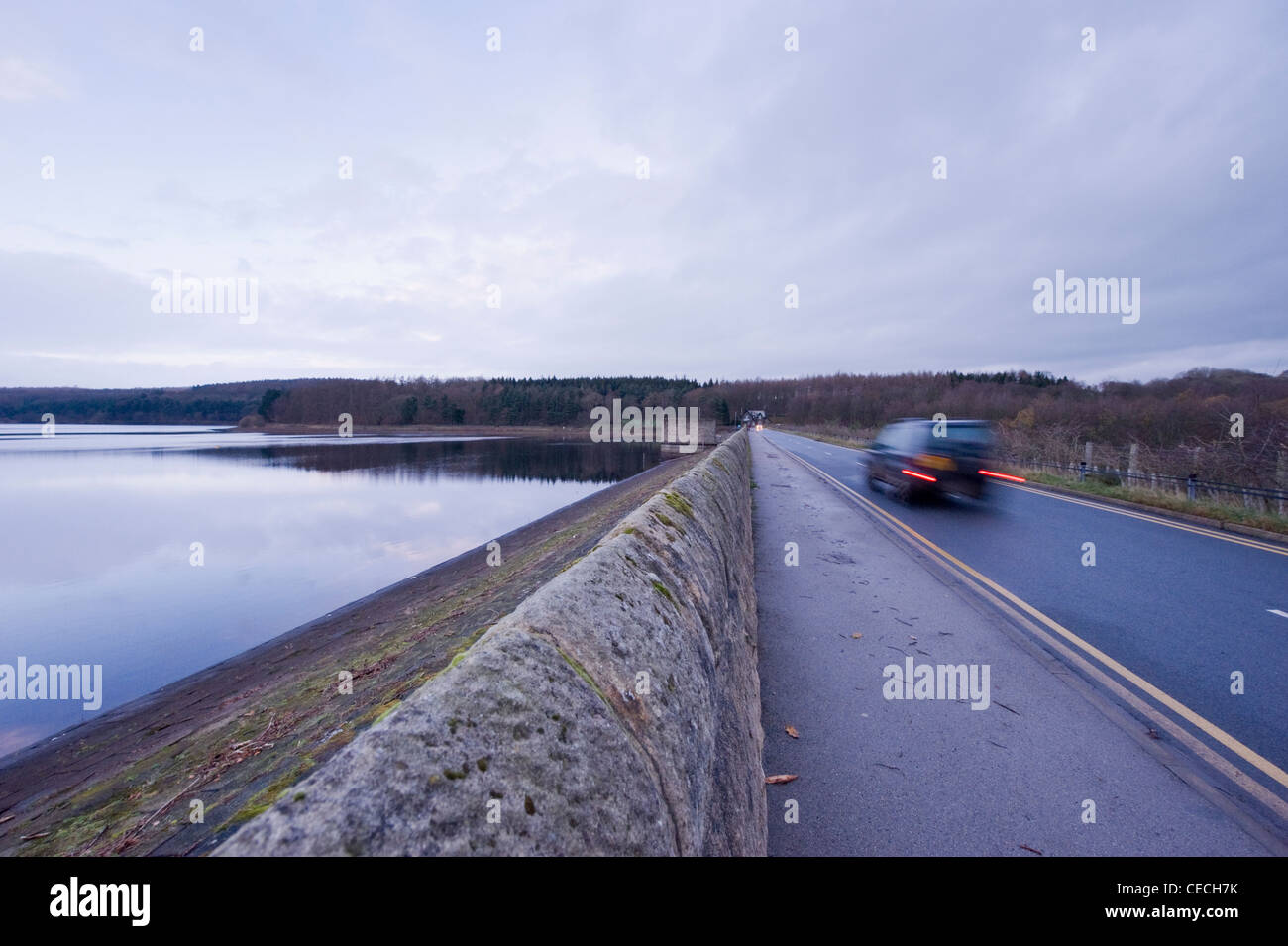 Car (4X4 vehicle + tail lights on) driving on road that crosses dam embankment by Fewston Reservoir (trees reflected in calm water) - Yorkshire, UK. Stock Photo