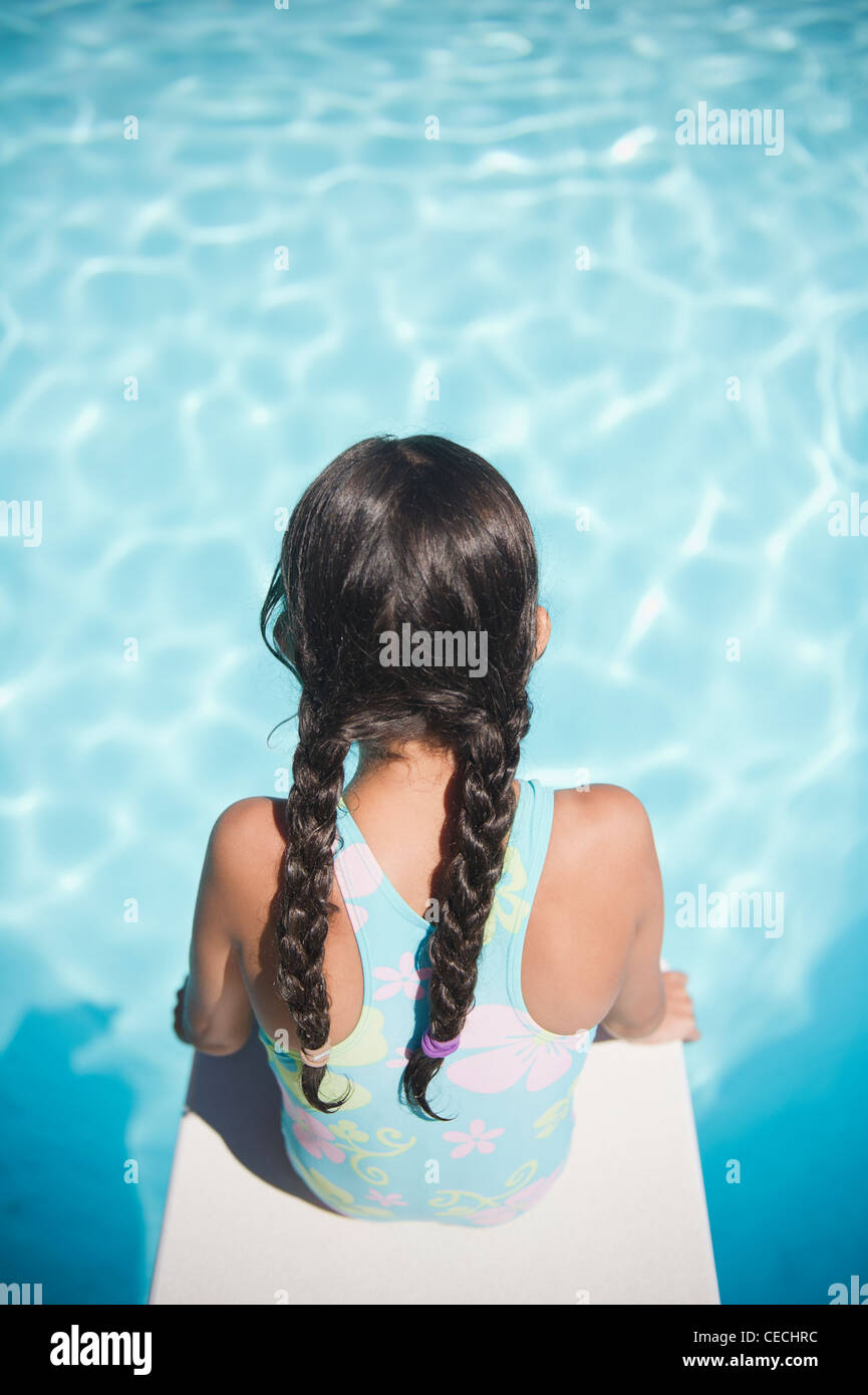 Hispanic Girl Sitting On Diving Board Over Swimming Pool Stock Photo