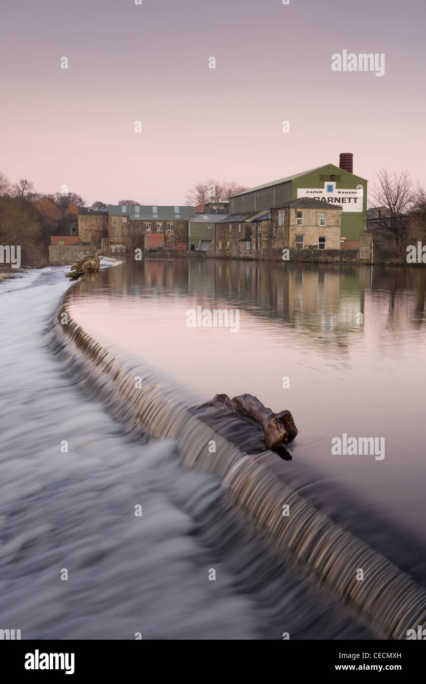 Flowing water of River Wharfe cascading over weir under pink sunset sky, historic Garnett's paper mill beyond - Otley, West Yorkshire, England, UK. Stock Photo