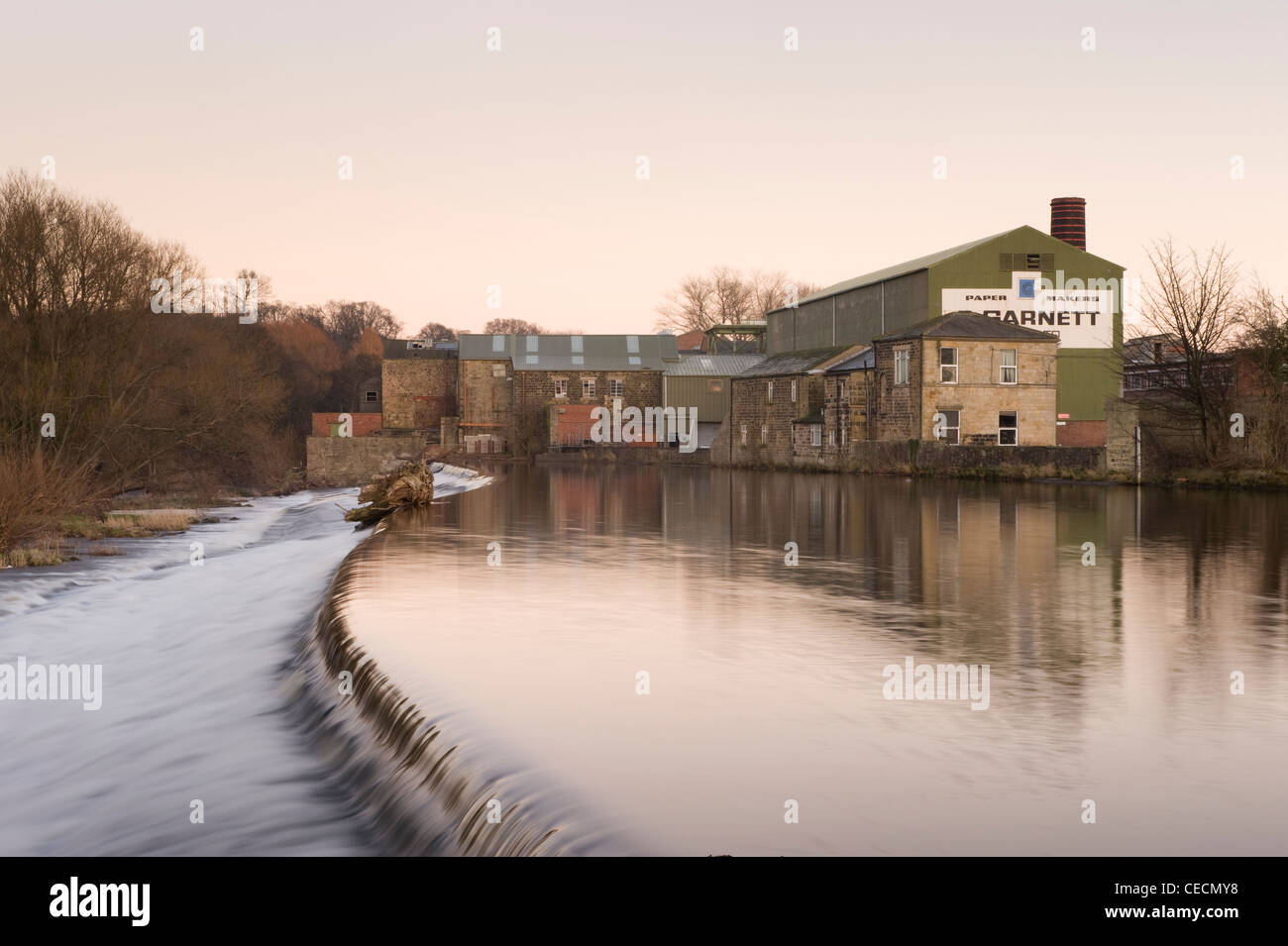 River Wharfe water gently flowing & cascading over weir under sunset sky, historic Garnett's paper mill beyond - Otley, West Yorkshire, England, UK. Stock Photo
