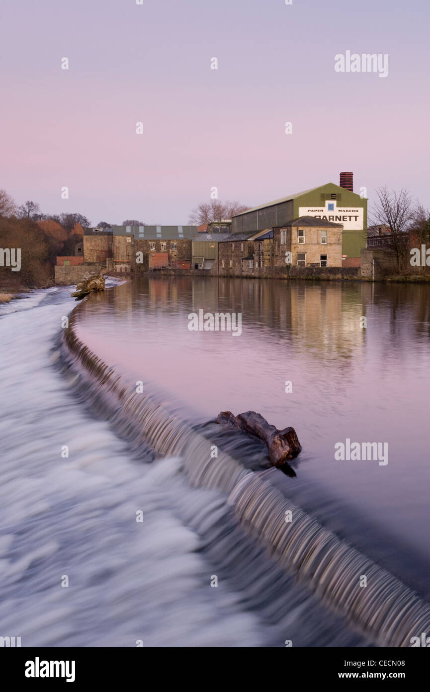 Flowing water of River Wharfe cascading over weir under pink sunset sky, historic Garnett's paper mill beyond - Otley, West Yorkshire, England, UK. Stock Photo
