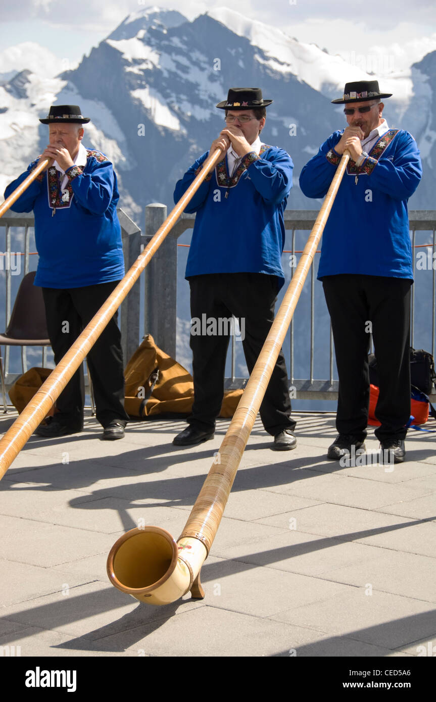 Horizontal close up portrait of three traditionally dressed Swiss men playing the Alpine horn with the Swiss Alps behind them. Stock Photo