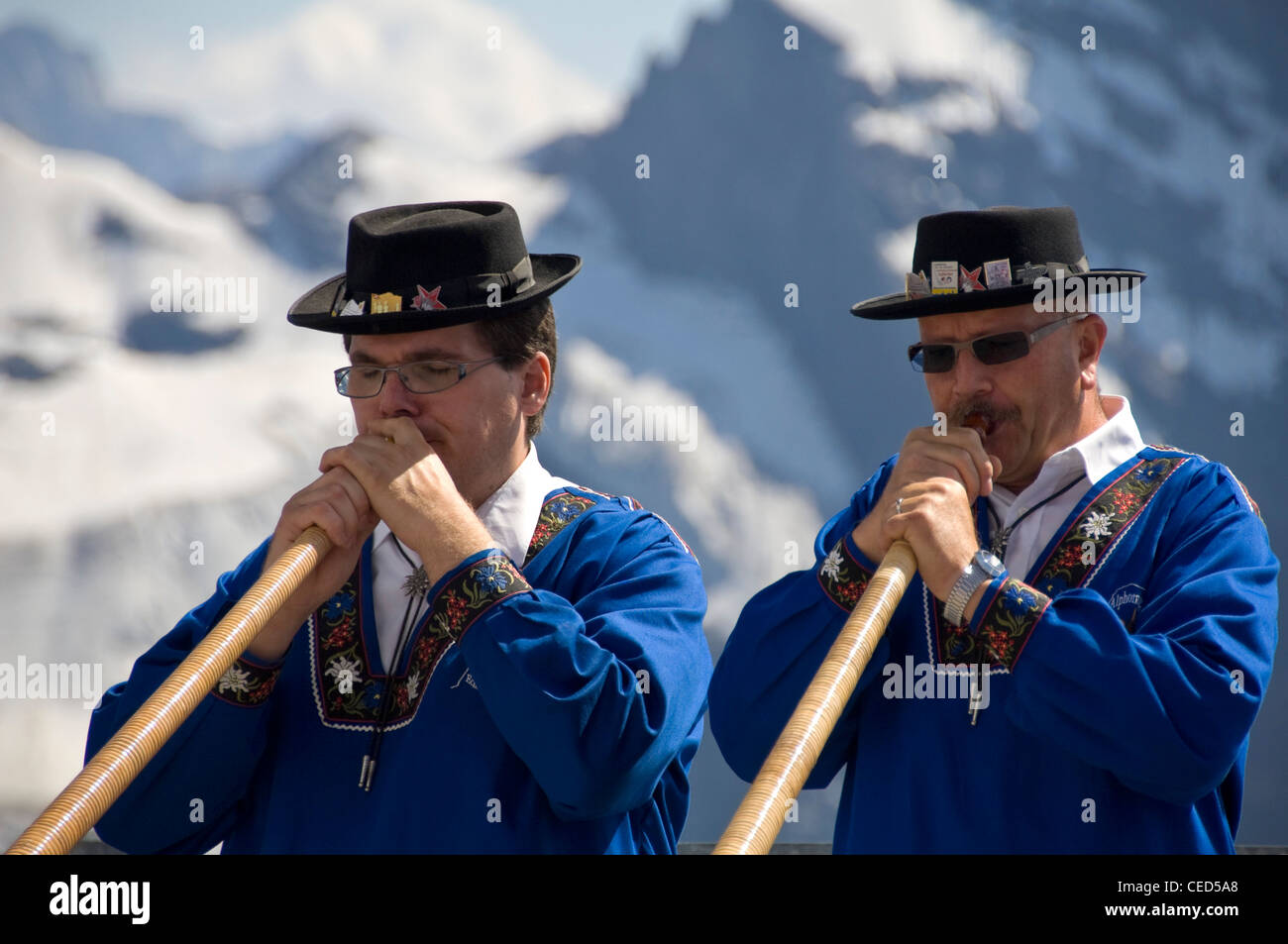Horizontal close up portrait of two traditionally dressed Swiss men playing the Alpine horn with the Swiss Alps behind them. Stock Photo